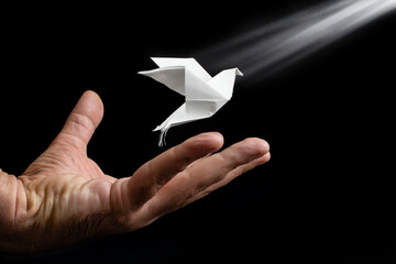 Male hand launching a floating white origami dove, peace offering or hope metaphor, on black background