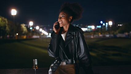 Cheerful lady talking cellphone sitting night street with champagne glass.