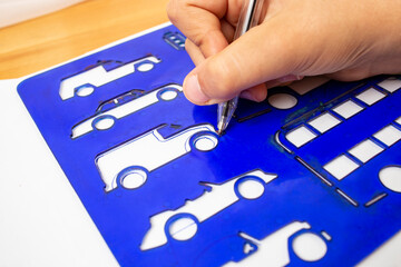 Child hand outlining and tracing a car shape with the help of a blue plastic stencil, soft focus close up, preschool leisure activity concept