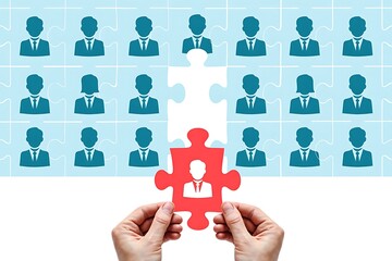 Hands holding a red puzzle piece with a person icon fitting into a larger puzzle of silhouetted business people representing recruitment and finding the right fit