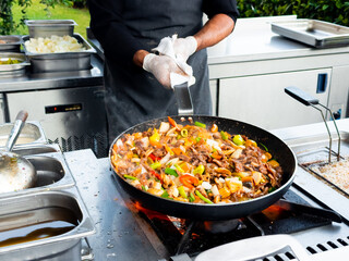 Male chef cooking stir fry outdoors with colorful vegetables and meat in a pan
