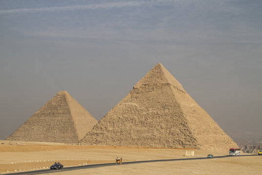 View of the ancient pyramids standing tall against the horizon, with vehicles traversing the dusty landscape in Giza, Al Jizah, Egypt.