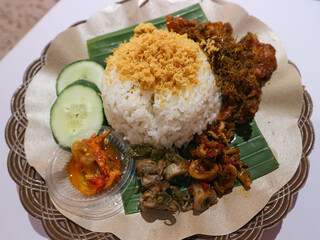 A traditional Indonesian rice dish served on a woven plate with various flavorful side dishes, including fried chicken, sambal, and squid