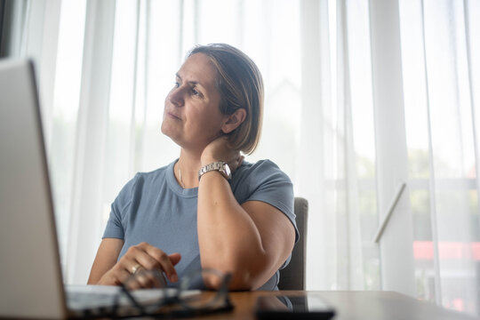 Woman experiencing neck pain while working on laptop at home, posture-related discomfort from computer desk job.	