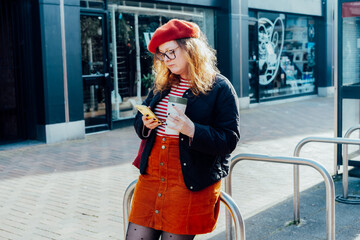 Young smiling plus size woman with backpack using mobile phone, drinking coffee outdoors on the street. Confident fashion hipster messaging, chatting, working remotely. Using navigator app for travel.
