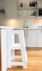 A minimalist white wooden toddler tower stands ready in a modern kitchen. It's designed to safely involve children in countertop activities and foster independence