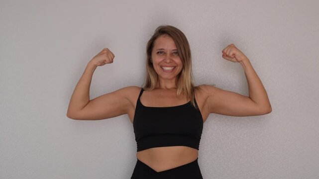 A smiling young blonde woman proudly flexes her biceps first lifting one arm and then the other on light grey background. A confident and joyful moment of self-expression, strength and body positivity