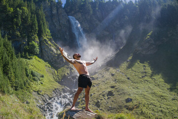 Carefree man in waterfall. Man freedom in waterfall landscape. Muscular man breathing fresh air on beauty waterfall. Topless model with raised hands on mountain nature landscape. Yoga by waterfall.