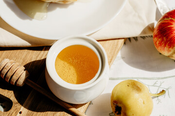 Cup of golden honey with dipper, fresh apples on rustic table, warm sunlight streaming through, cozy autumn atmosphere.