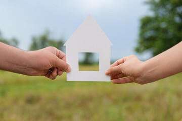 Two hands hold a white paper cutout of a house in a grassy field, symbolizing dreams of building a home, connection, and new beginnings.