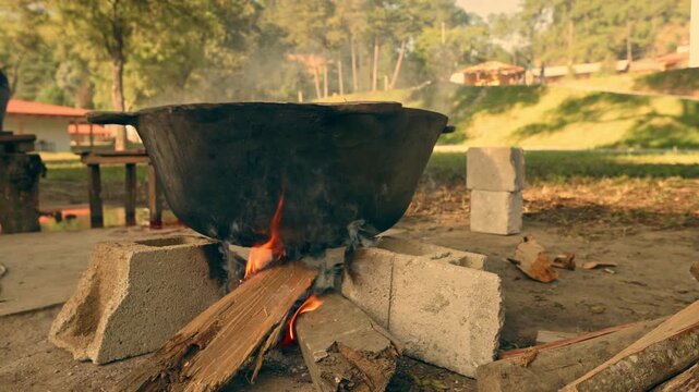 An industrial cauldron of boiling water sits on a fire on the ground. A scene in the middle of a field surrounded by vegetation.