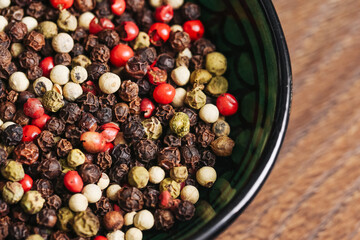 Colorful mix of peppercorns in ceramic bowl. Red, black, white and green dried pepper seeds closeup. Spicy cooking ingredients on wooden kitchen table. Natural food seasoning. Organic culinary spice.