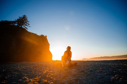 Brunette woman wearing a yellow rain jacket on Ruby Beach sitting at sunset. Scenic coastal view with sea stacks, tidepools, and warm twilight colors. Olympic National Park, Washington State.