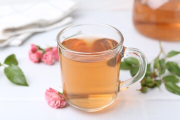 Aromatic tea in glass cup, roses and green leaves on white table, closeup