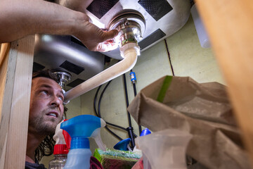 A man uses a flashlight to inspect or repair plumbing beneath a kitchen sink, surrounded by household cleaning supplies. This candid, real-life scene highlights home maintenance, DIY repairs, and resp