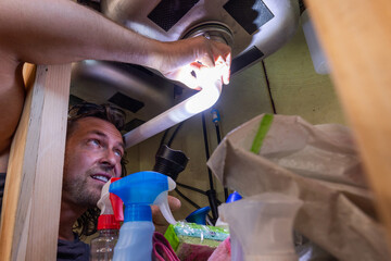 A man uses a flashlight to inspect or repair plumbing beneath a kitchen sink, surrounded by household cleaning supplies. This candid, real-life scene highlights home maintenance, DIY repairs, and resp