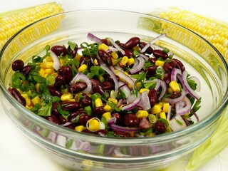 Glass bowl with vegetable salad based on corn,  corn cobs are on a white table. Ingredients, fresh corn on the cob, red bean, red onion, dill, parsley and green scallions