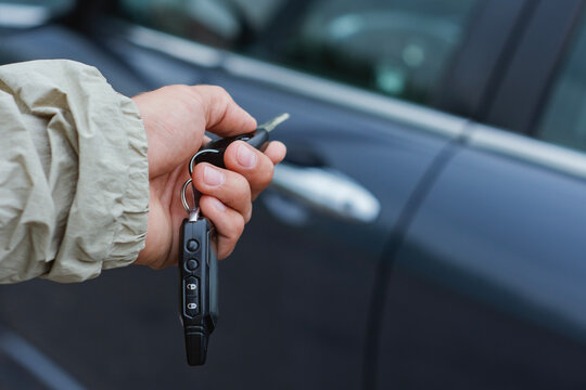 Close-up of a hand holding car keys and a key fob with remote control buttons against the background of a car. Concept of car ownership and rental.