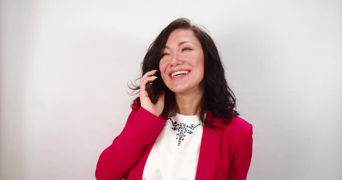 Young woman in red blazer smiling and laughing while talking on the phone in front of a light grey background space