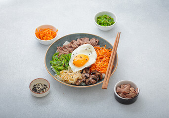 Bibimbap, authentic korean dish with a steamed rice topped with fried egg, beef, colorful vegetables, traditional dish of Korea on light background with chopsticks