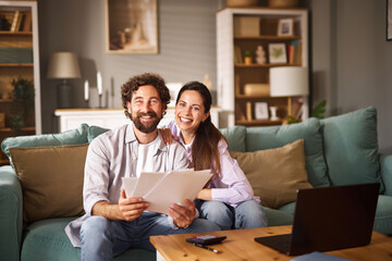 A joyful couple sits on a comfortable couch, smiling as they examine life insurance papers. Their...