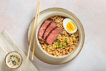 Bowl filled with ramen noodles, tender sliced beef, boiled egg and fresh green onions, asian style soup on light beige background top view