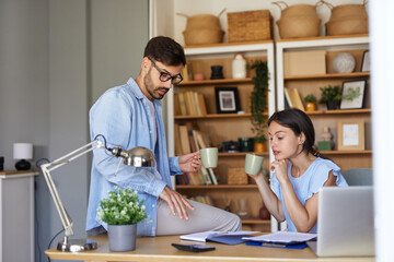 Obraz premium Two colleagues engage in a discussion at a home office, sipping tea from mugs while surrounded by books and plants, creating a productive yet relaxed atmosphere.