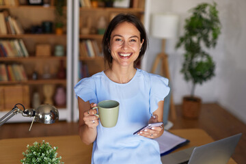 A woman stands in her home office, smiling while holding a mug in one hand and a smartphone in the other. The warm atmosphere invites productivity. © Stockphotodirectors
