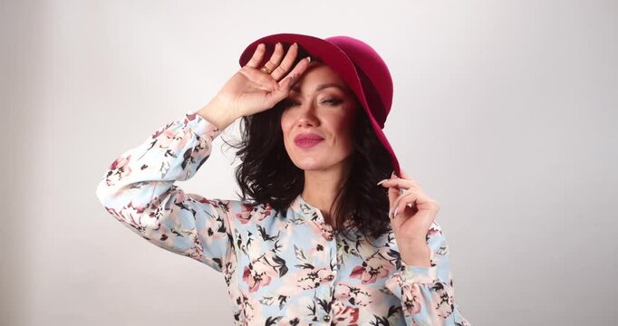 Woman in front view, close to camera, removes her hat with one hand while gazing directly into lens. Captured in medium close-up, emphasizing her expression and gesture.