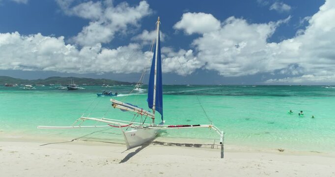 Traditional paraw sailboat with a blue sail resting on the shore of White Beach in Boracay, Philippines, surrounded by turquoise water and distant sailing boats