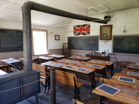Interior of a historical schoolhouse with wooden desks, slate boards, chalkboards, and a central iron stove. Union Jack flag and moral lessons on the wall 