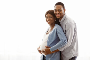 Obraz premium Smiling black man hugging his pregnant cheerful woman over white background, panorama with copy space. Happy african american expecting couple in comfortable clothes posing at home