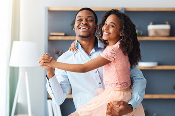 Cheerful little black girl in beautiful dress dancing with her dad at home, african american father...