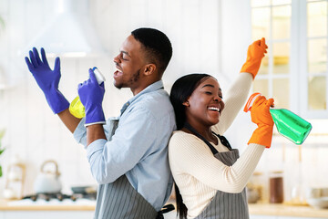 Joyful black couple using spray bottles as mics, singing during cleanup at kitchen. Happy young...