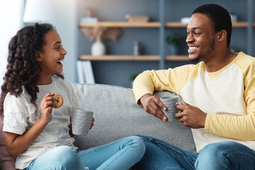 Cute black father and daughter drinking tea with cookies in living room, sitting on couch and...