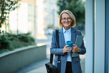 Portrait of a middle aged mature businesswoman woman using a laptop computer and holding a cup of coffee  in a city park, surrounded by modern corporate office buildings