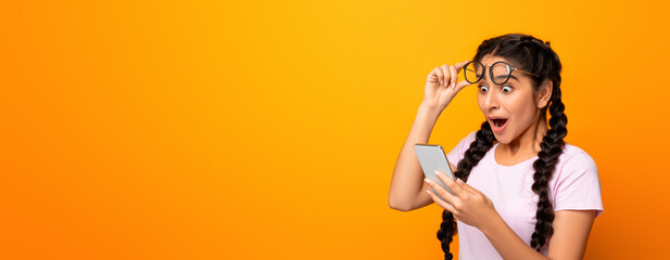 Obraz premium Wow, Great News. Portrait Of Excited Surprised Indian Woman Holding And Using Smartphone, Browsing Social Media. Emotional Young Lady Having Success, Taking Off Glasses, Yellow Studio Background