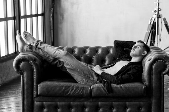Black and white portrait of strong healthy handsome sexy athletic man in a shirt with naked torso lying on a leather sofa in a stylish loft interior. Male model posing and smiling