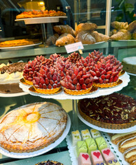 A vibrant bakery window display filled with an irresistible assortment of tarts and cakes, adorned with fresh strawberries and luscious creamy toppings. The colorful, inviting arrangement captures the