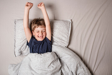 Smiling boy waking up joyfully, stretching arms in bed. Positive morning vibes, happy childhood and energy of a new day. Good sleep and fresh start. Child having fun before or after healthy sleeping. 