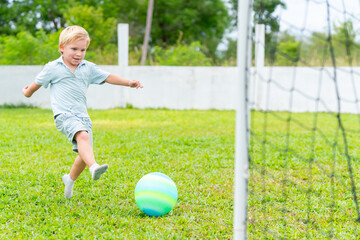 Happy little Caucasian boy playing football on grass field playground in summer. Kindergarten children enjoy and fun outdoor active lifestyle playing outside classroom and exercising at kid area.