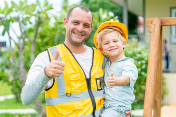 Naklejka premium Portrait of Happy Caucasian family father and little son in safety jacket and hard hat standing in front of the house. Labor day, father's day, construction engineering and safety occupation concept.