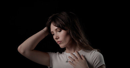 Young woman with hand in hair looking down wearing white t shirt and silver necklace on black background