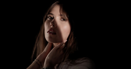 Naklejka premium Beautiful young woman with light makeup is posing in a studio with her hands on her neck, illuminated by a soft light