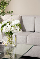 Close-Up of Delicate White Roses with Fresh Green Leaves in a Transparent Glass Vase, Set on a Glass Coffee Table, Glimpse of Textured Fabric Light Grey Sofa in the Backdrop, Soft Natural Light.