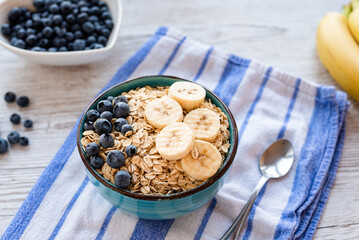 Porridge oatmeal with fresh fruit, a healthy breakfast bowl with oats, banana slices, and blueberries on a striped kitchen towel and rustic wooden table, with a spoon on the side.
