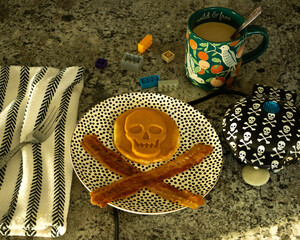 A whimsical flatlay of a skull-and-crossbones pancake breakfast with crispy bacon on a patterned plate. © Brandi