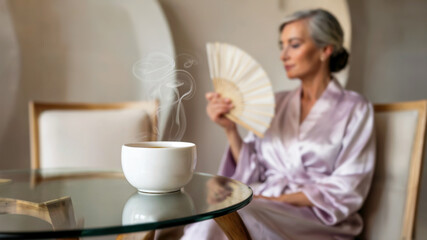 White tea cup with herbal drink on glass table, elegant elderly woman in robe holding a fan in the background. Concept of menopause and calm self-care