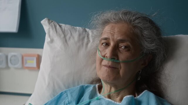 Close up of silver haired woman turning her head and looking at camera gloomily while lying in hospital bed in intensive care ward