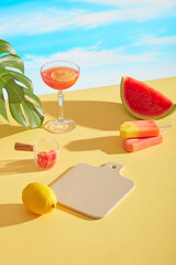 Vibrant summer setup with cocktails, citrus fruits, watermelon slices, popsicle, and tropical leaves on yellow surface under sunny blue sky, ideal for cosmetics or SPF mockups.